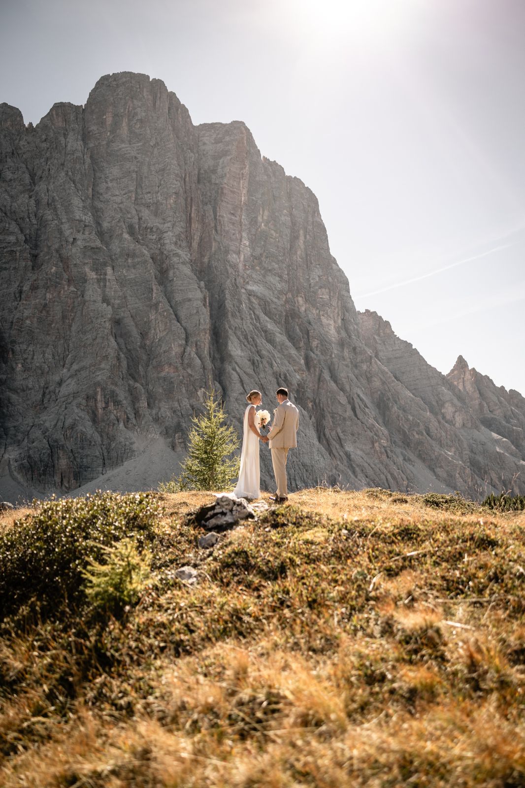 Ein Hochzeitspaar hält sich vor einer sonnigen Bergkulisse an den Händen.